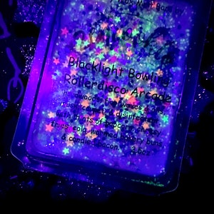 Puede incluir: Una barra de cera de soja de 2,5 onzas con aroma a bolos de luz negra y roller disco. La cera fundida está en un recipiente cuadrado transparente con borde negro. El recipiente está lleno de purpurina de colores y estrellas que brillan bajo una luz negra.