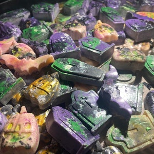 Puede incluir: Una colección de fundentes de cera con temática de Halloween en varias formas, incluyendo murciélagos, calabazas y lápidas. Los fundentes están decorados con purpurina verde, morada y dorada y tienen un aspecto moteado.
