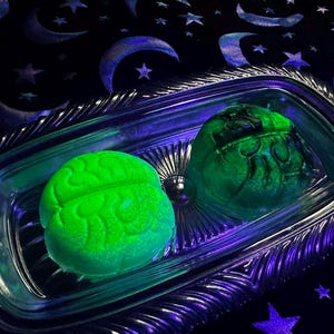Könnte beinhalten: Zwei grüne, gehirnförmige Badekugeln in einer durchsichtigen Glasschale mit silberfarbenem, verziertem Rand. Der Hintergrund ist ein dunkelblauer Stoff mit Monden und Sternen.