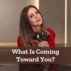 May include: A set of tarot cards featuring dark backgrounds with colorful celestial and cosmic illustrations, held in a fan shape. The cards display intricate gold-toned details and vibrant patterns. The text "What Is Coming Toward You?" is visible.