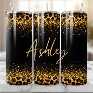 May include: A personalized tumbler with a black background, gold glitter accents, and a leopard print design. The name "Ashley" is written in gold script across the center. The tumbler includes a straw.
