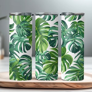 Puede incluir: Tres vasos de acero inoxidable con un diseño de hojas tropicales. Los vasos son blancos con hojas verdes y tienen una tapa plateada.