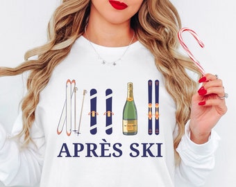 Op de afbeelding: Witte longsleeve trui met een afbeelding van ski-stokken, een fles champagne en de tekst "APRÈS SKI" in blauw.