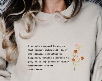 Puede incluir: Una sudadera beige con una cita de Jane Austen impresa en la parte delantera. La cita dice: "Solo estoy decidida a actuar de esa manera, que, en mi opinión, constituye mi felicidad, sin referencia a ti, ni a ninguna persona tan completamente desconectada de mí." La cita está rodeada de un pequeño diseño floral.