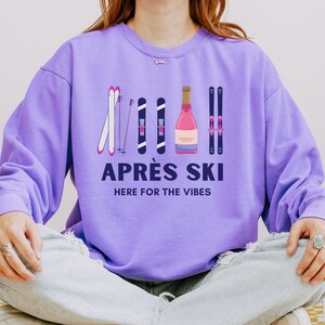 May include: Purple sweatshirt with a graphic of ski poles and a bottle of champagne. The text "APR&Egrave;S SKI HERE FOR THE VIBES" is printed on the sweatshirt.