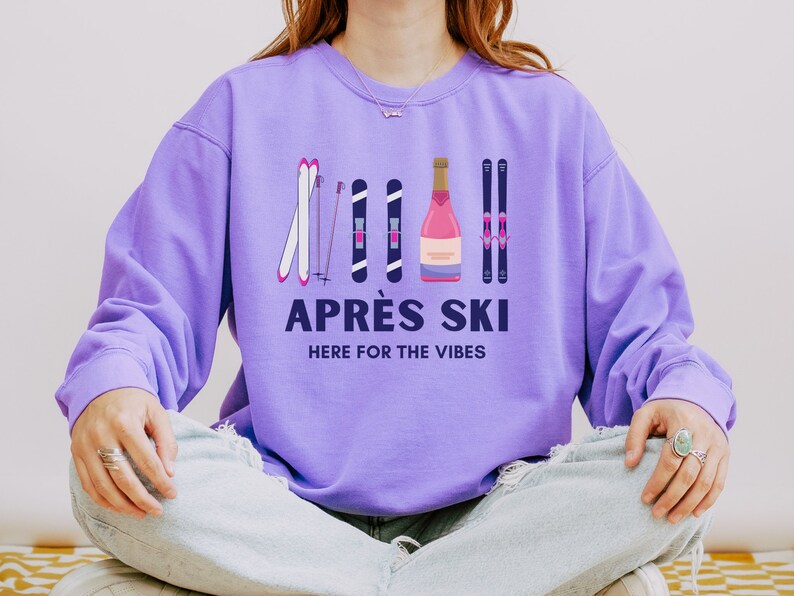 May include: Purple sweatshirt with a graphic of ski poles and a bottle of champagne. The text "APR&Egrave;S SKI HERE FOR THE VIBES" is printed on the sweatshirt.