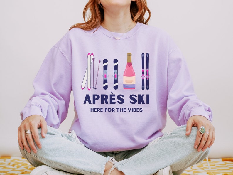 May include: A lavender sweatshirt with a graphic design of ski poles, skis, and a bottle of champagne. The text "APR&Egrave;S SKI HERE FOR THE VIBES" is printed on the sweatshirt.