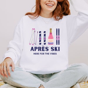 May include: White sweatshirt with a graphic design of pink ski poles and a bottle of pink champagne. The text "APR&Eacute;S SKI HERE FOR THE VIBES" is printed in blue below the graphic.