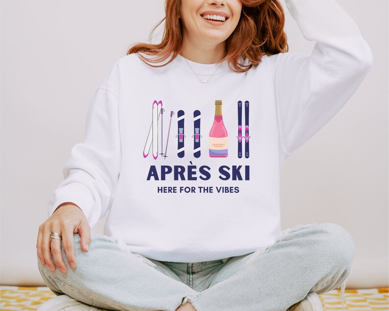 May include: White sweatshirt with a graphic design of pink ski poles and a bottle of pink champagne. The text "APR&Eacute;S SKI HERE FOR THE VIBES" is printed in blue below the graphic.