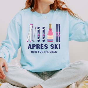 May include: A light blue sweatshirt with a graphic design of ski poles, skis, and a bottle of champagne. The text "APR&Egrave;S SKI HERE FOR THE VIBES" is printed below the graphic.