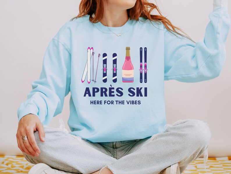 May include: A light blue sweatshirt with a graphic design of ski poles, skis, and a bottle of champagne. The text "APR&Egrave;S SKI HERE FOR THE VIBES" is printed below the graphic.