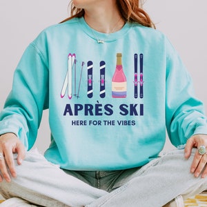 May include: A turquoise blue sweatshirt with a graphic design of ski poles, skis, and a bottle of champagne. The text "APR&Eacute;S SKI HERE FOR THE VIBES" is printed on the sweatshirt.