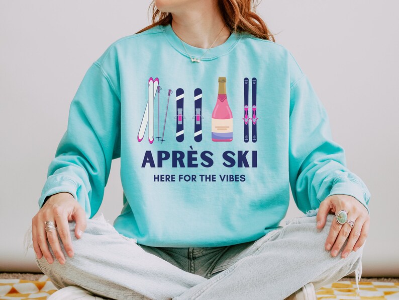 May include: A turquoise blue sweatshirt with a graphic design of ski poles, skis, and a bottle of champagne. The text "APR&Eacute;S SKI HERE FOR THE VIBES" is printed on the sweatshirt.