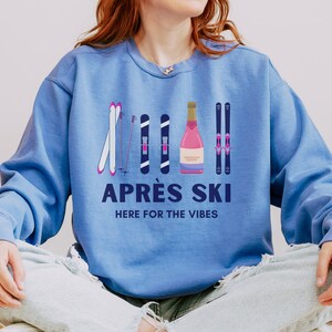 May include: A blue sweatshirt with a graphic design of ski poles, skis, and a bottle of champagne. The text "APR&Eacute;S SKI HERE FOR THE VIBES" is printed on the sweatshirt.