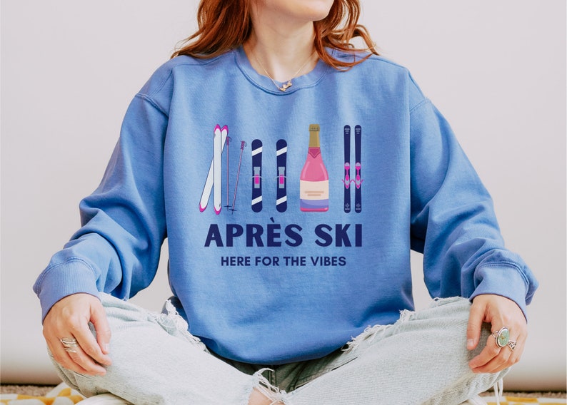 May include: A blue sweatshirt with a graphic design of ski poles, skis, and a bottle of champagne. The text "APR&Eacute;S SKI HERE FOR THE VIBES" is printed on the sweatshirt.