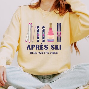 May include: A yellow sweatshirt with a graphic design of skis, ski poles, and a bottle of champagne. The text "Apr&eacute;s Ski" and "Here for the Vibes" is printed on the sweatshirt.