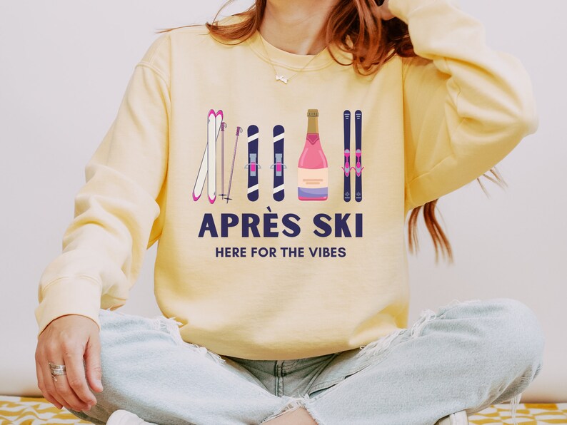 May include: A yellow sweatshirt with a graphic design of skis, ski poles, and a bottle of champagne. The text "Apr&eacute;s Ski" and "Here for the Vibes" is printed on the sweatshirt.