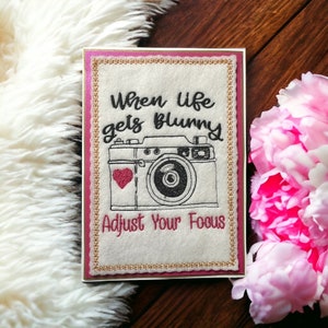 May include: A white card with a pink border and gold trim features a black and white illustration of a vintage camera with a red heart. The text "When life gets blurry Adjust your focus" is embroidered in black on the card.