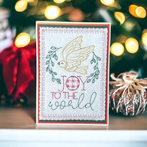 May include: A handmade Christmas card with a gold embroidered dove and the words "JOY TO THE WORLD." The card has a red and green border and is set against a blurred background with Christmas decorations.