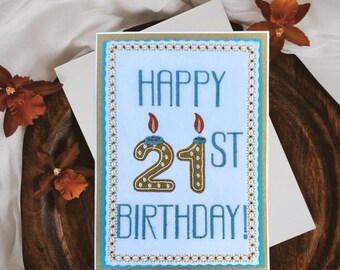 May include: A white card with a blue and gold border. The card says "Happy 21st Birthday!" with the number 21 in gold with candles on top.