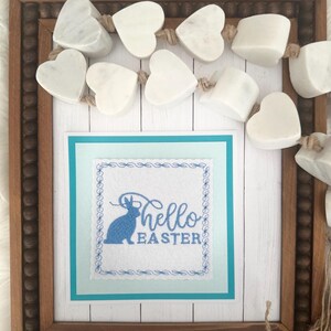 May include: A framed embroidery design with a blue bunny and the text "hello easter" on a white background. The frame is decorated with a garland of white heart-shaped beads.