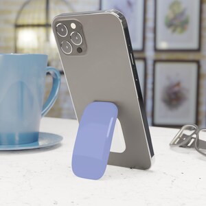 May include: A gray smartphone propped up by a smooth, light purple oval-shaped adhesive phone stand attached to the back. The device features a triple-lens camera system on the upper left corner and a metallic frame, resting at an angled position.