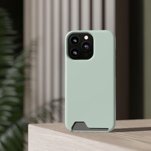 May include: A light sage green smartphone case with a smooth matte finish. The case features a rectangular cutout at the bottom revealing a dark gray interior layer and a square camera module cutout at the top left with three circular lenses.