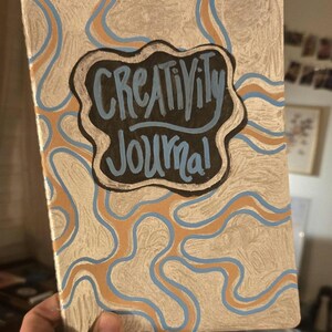 May include: A hand-painted journal with a silver background and wavy orange and blue lines. The journal cover features the words "Creativity Journal" in a black and blue design.