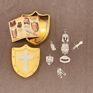May include: A gold-colored metal box shaped like a shield with a white cross inside. The box is open and reveals a small booklet with the text "The Armor of God" and illustrations of different pieces of armor. There are also several small silver charms, including a sword, a shield, a helmet, and a breastplate.