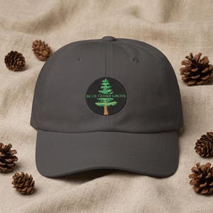 May include: A charcoal gray baseball cap featuring a circular embroidered patch on the front. The patch displays a green cedar tree with a brown trunk and the text BLUE CEDAR GROVE in green lettering centered over the tree graphic.