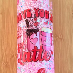 Puede incluir: Vaso rosa con purpurina y un gráfico rojo que dice "Love you a latte" con imágenes de bebidas de café y corazones.