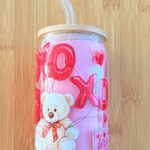 Puede incluir: Un vaso de vidrio transparente con fondo rosa y un diseño de osito de peluche. El vaso tiene una tapa de bambú y una pajita de vidrio transparente. El diseño presenta globos rojos que deletrean "XO" y corazones. El vaso también tiene una etiqueta rosa que dice "Love".