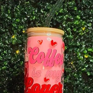 Puede incluir: Un vaso de vidrio rosa con tapa transparente y una pajita. El vaso tiene las palabras "Coffee is my lover" escritas en letras rosa y rojas. Hay pequeños corazones rosas en el vaso.