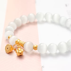 May include: A white gemstone bracelet with gold accents. The bracelet is made of round, white beads and has a gold charm with a floral design.