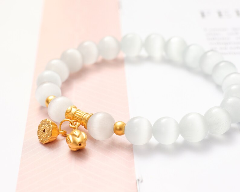 May include: A white gemstone bracelet with gold accents. The bracelet is made of round, white beads and has a gold charm with a floral design.