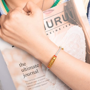 May include: A white braided bracelet with a gold charm that says "LOVE" in gold letters. The bracelet is on a person's wrist.