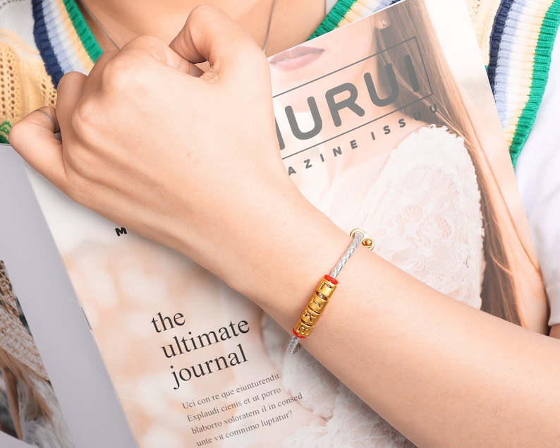 May include: A white braided bracelet with a gold charm that says "LOVE" in gold letters. The bracelet is on a person's wrist.