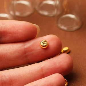 May include: Close-up of small, gold-colored, round beads. Each bead has a small, embossed design. The beads are resting on a brown surface. The image is well-lit, highlighting the details of the beads.