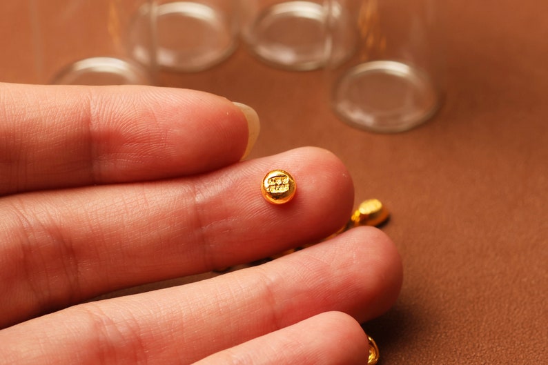 May include: Close-up of small, gold-colored, round beads. Each bead has a small, embossed design. The beads are resting on a brown surface. The image is well-lit, highlighting the details of the beads.