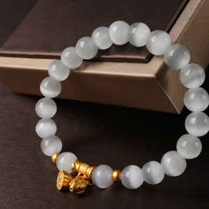May include: A white gemstone bracelet with a gold charm. The bracelet is made of round beads and has a gold clasp.