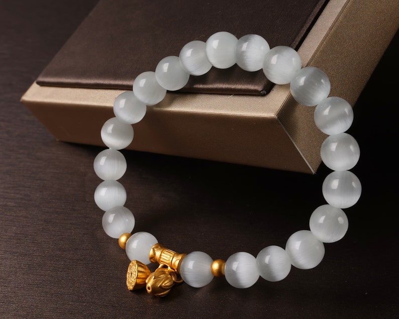 May include: A white gemstone bracelet with a gold charm. The bracelet is made of round beads and has a gold clasp.