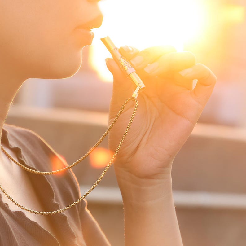 May include: A gold chain necklace with a gold whistle pendant. The whistle is being held by a person's hand.