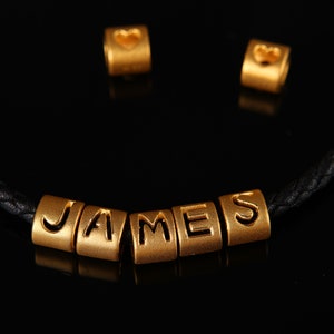 May include: A black leather bracelet with gold beads spelling out the name "JAMES". Two additional gold beads with heart shapes are also included.