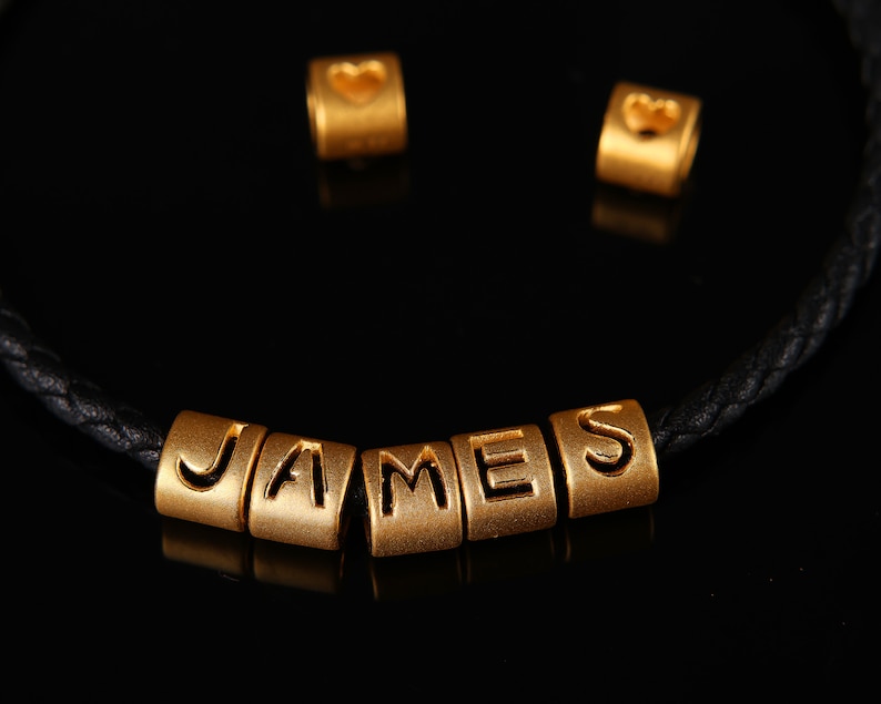 May include: A black leather bracelet with gold beads spelling out the name "JAMES". Two additional gold beads with heart shapes are also included.