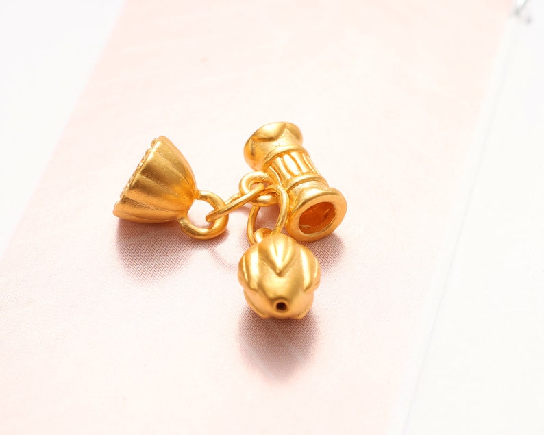 May include: Gold-toned metal charm with a flower-shaped design and a cylindrical bead with a decorative pattern.