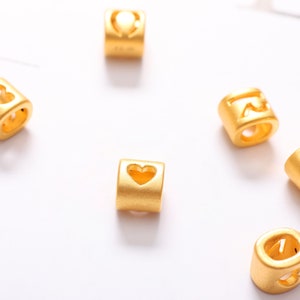 May include: Gold-colored metal beads with a heart-shaped cutout. The beads are designed for use in jewelry making.