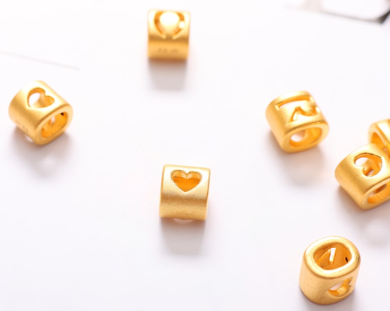 May include: Gold-colored metal beads with a heart-shaped cutout. The beads are designed for use in jewelry making.