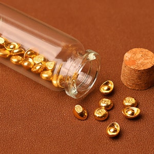 May include: A clear glass vial tipped on its side, spilling out small, gold-colored ingots. The vial's cork stopper sits nearby. The ingots are scattered on a brown surface, reflecting light.