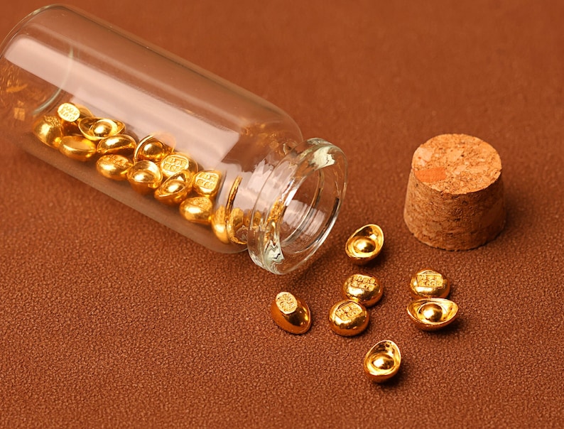 May include: A clear glass vial tipped on its side, spilling out small, gold-colored ingots. The vial's cork stopper sits nearby. The ingots are scattered on a brown surface, reflecting light.