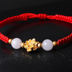 May include: A red string bracelet with two white beads and a gold lotus flower charm.